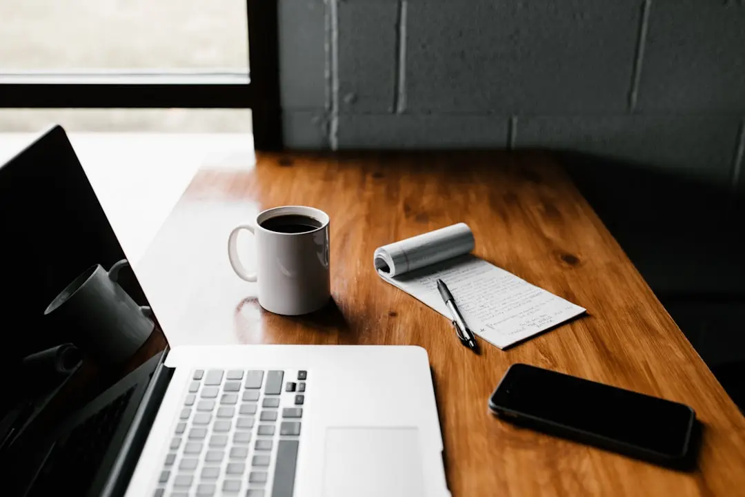 A cozy workspace featuring a laptop, a white coffee mug, a notepad with handwritten notes, a pen, and a smartphone on a wooden desk near a window.